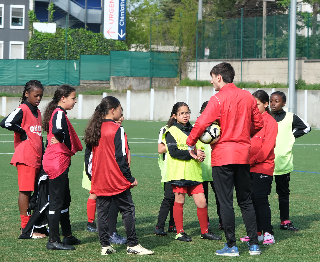 Un groupe de jeunes filles en tenue de sport sur un terrain de football écoutent un entraineur
